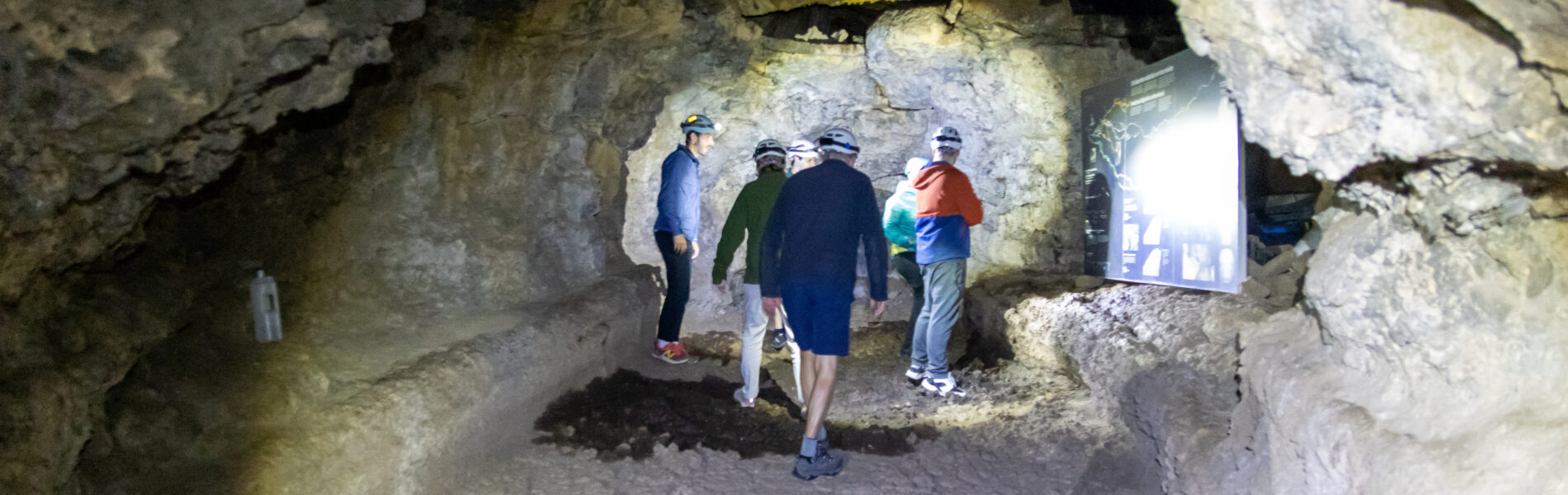 Visitors walking through a wide lava tube with pale tan stone walls and ceiling, bright daylight entering from an opening on the right.