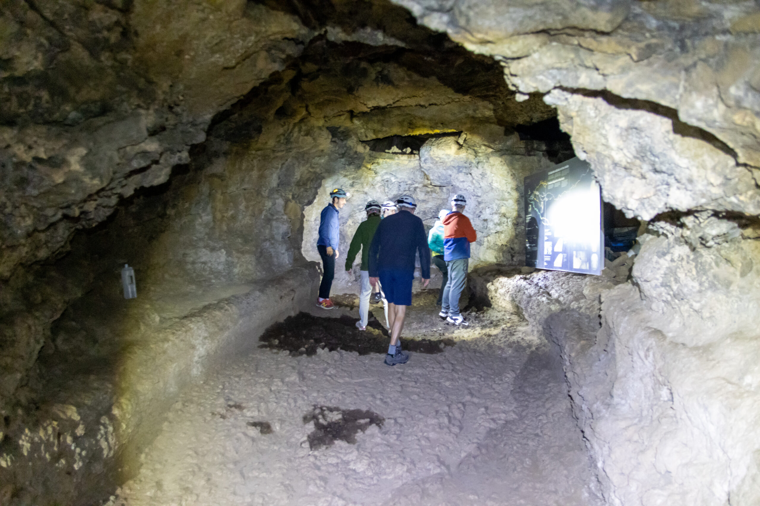 Visitors walking through a wide lava tube with pale tan stone walls and ceiling, bright daylight entering from an opening on the right.