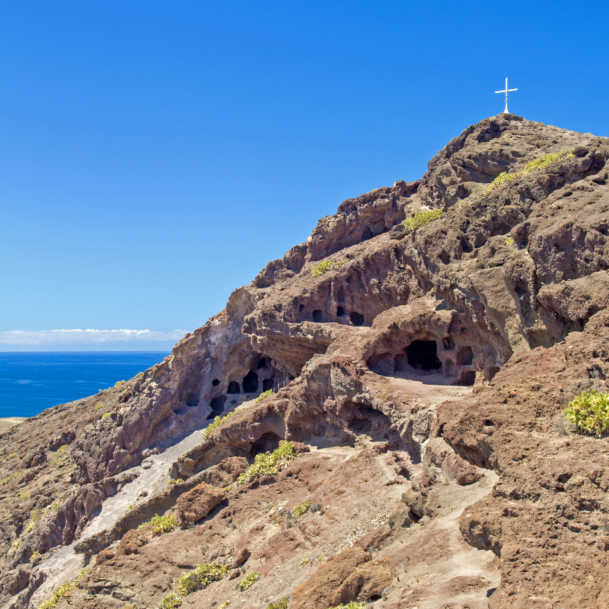 A white cross crowns the volcanic ridge above El Confital, where ancient caves pierce the rust‑coloured rock face.