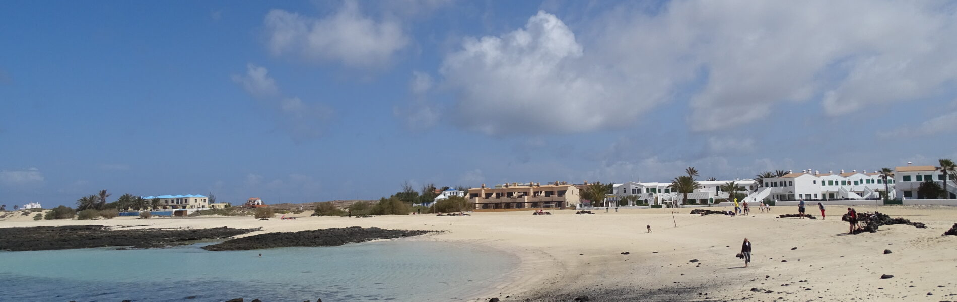 Sandy beach with turquoise shallow water, dark volcanic rocks in foreground, whitewashed buildings and palm trees along the shore under blue sky with white clouds
