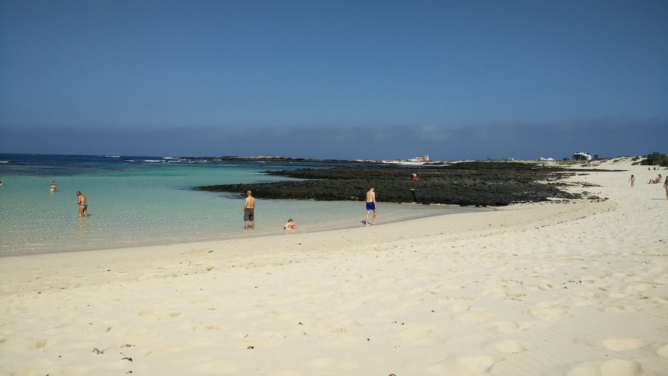 El Cotillo Beach features pristine turquoise waters, soft sand, and distinctive dark volcanic rock formations, with calm conditions ideal for swimming and wading.