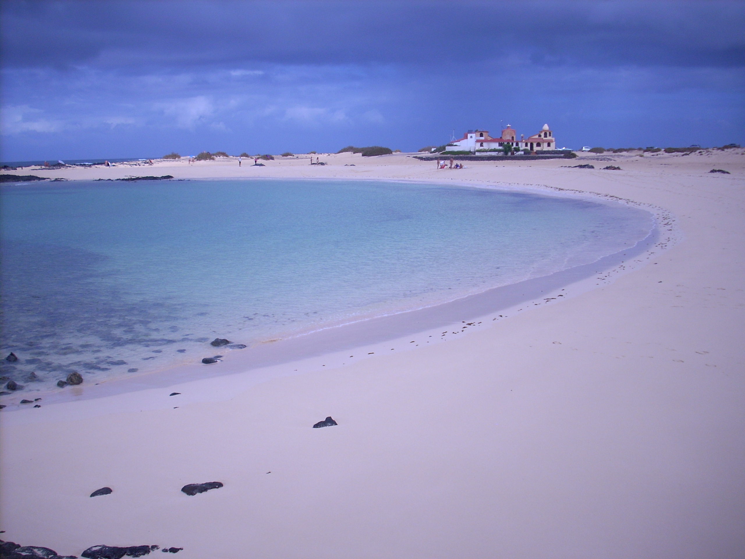 El Cotillo Beach offers a sheltered crescent cove with calm turquoise waters and pristine sand, backed by the distinctive white and terracotta lighthouse buildings.