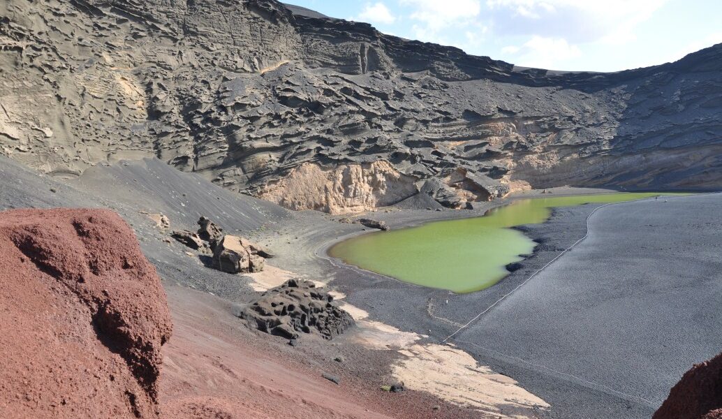 Volcanic crater lake with distinctive bright green water surrounded by dark lava fields, red cinder cones, and layered basalt cliffs under blue sky