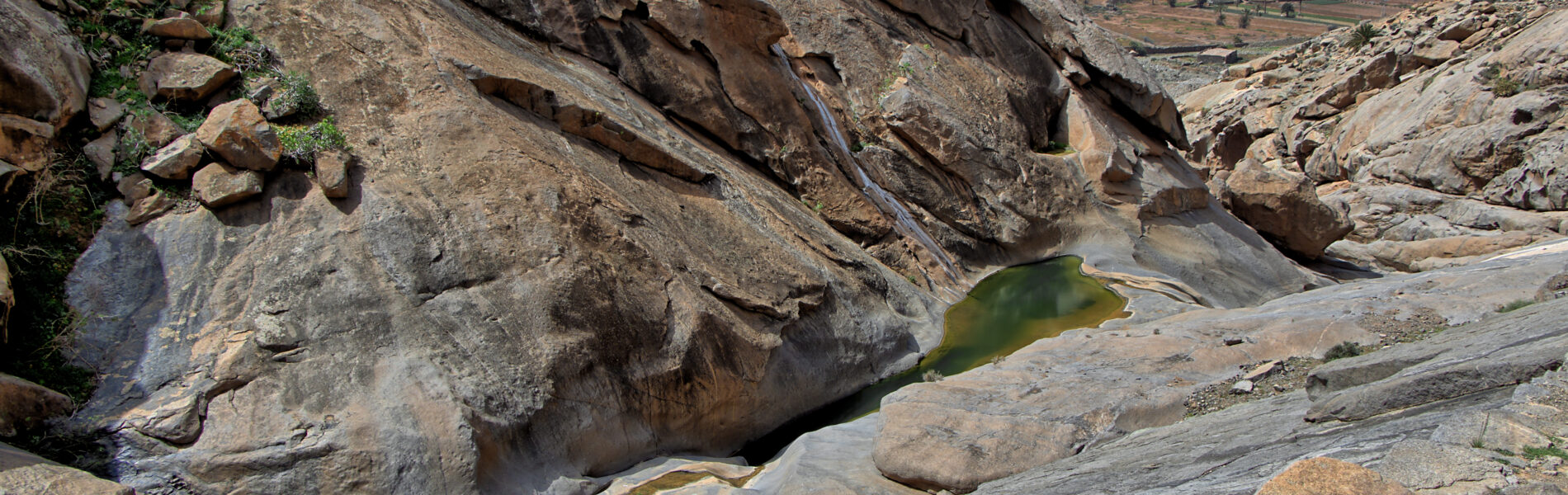 Rocky canyon with clear turquoise-green pools of water flowing between weathered stone formations, arid mountains and scattered buildings visible in distant valley