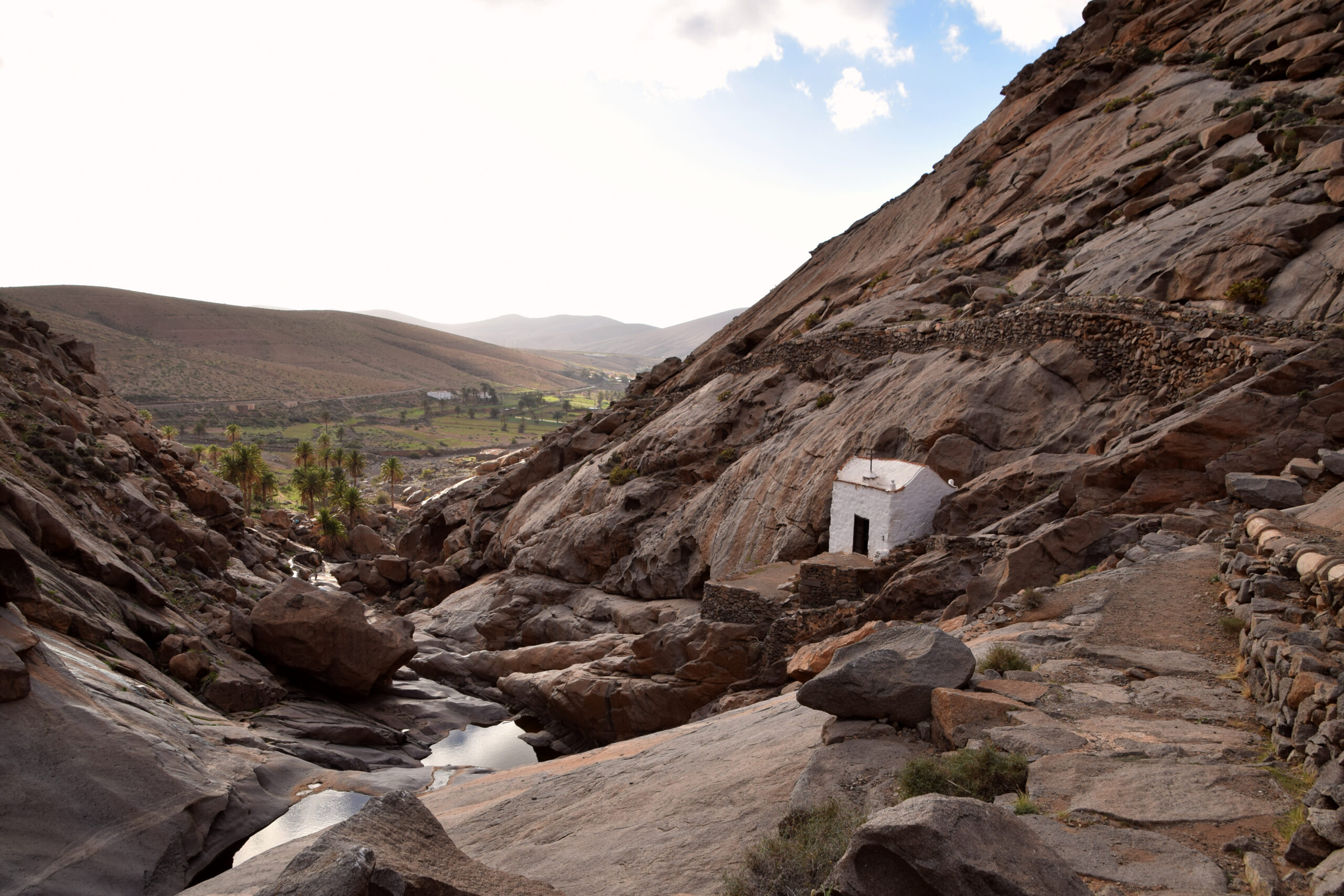 A traditional white structure shelters in the rocky canyon landscape near Embalse de las Peñitas, with cultivated valley lands stretching toward distant mountains.