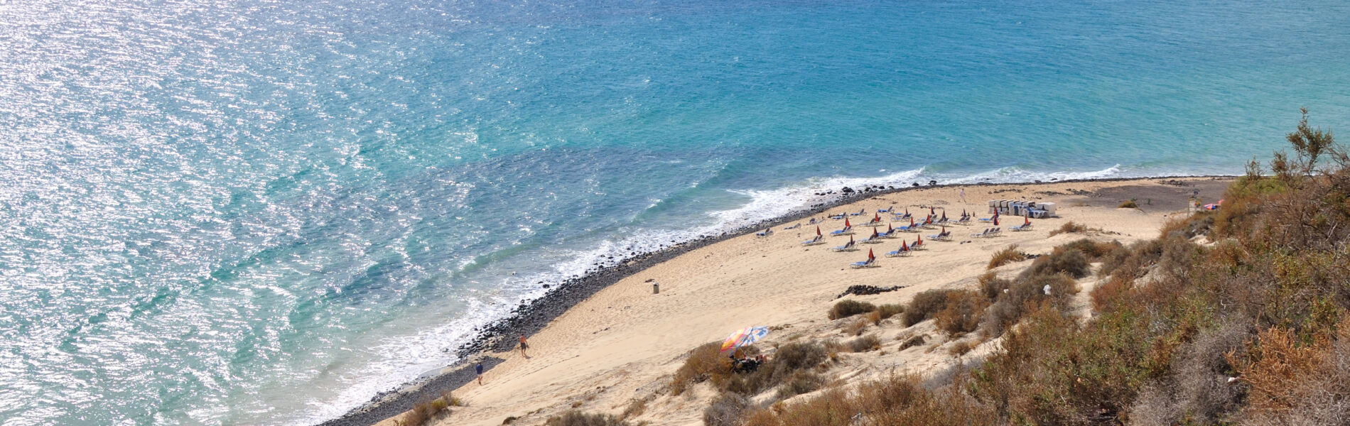 Aerial view of a sandy beach with turquoise water, white-capped waves, scattered beachgoers, and sparse dry vegetation on the surrounding cliffs