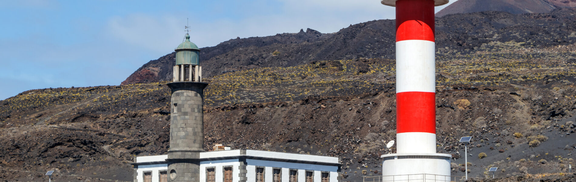 Red and white striped lighthouse with white keeper's building beside a darker stone tower, set against dark volcanic slopes.