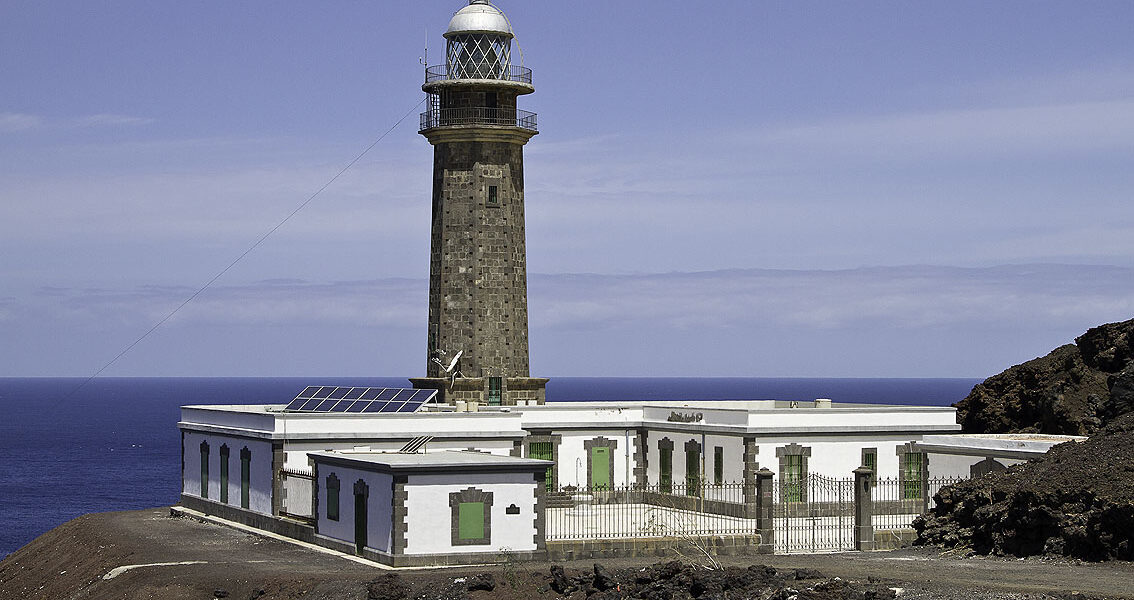 Stone lighthouse tower with white lantern room and cross-topped cupola, adjacent to a white keeper's house with green shutters on volcanic black rock shoreline facing blue ocean.