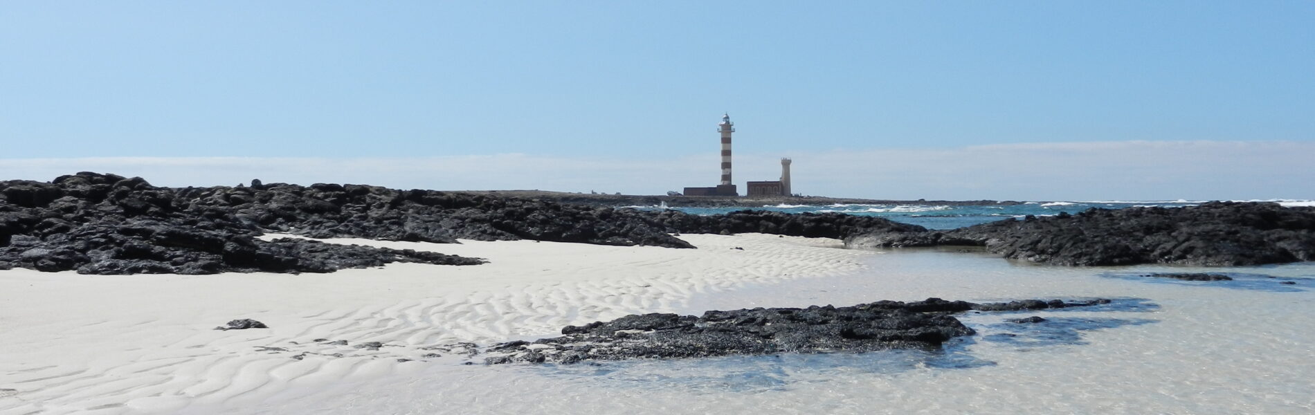 Striped lighthouse and ruined tower on rocky outcrop across shallow turquoise water from sandy beach