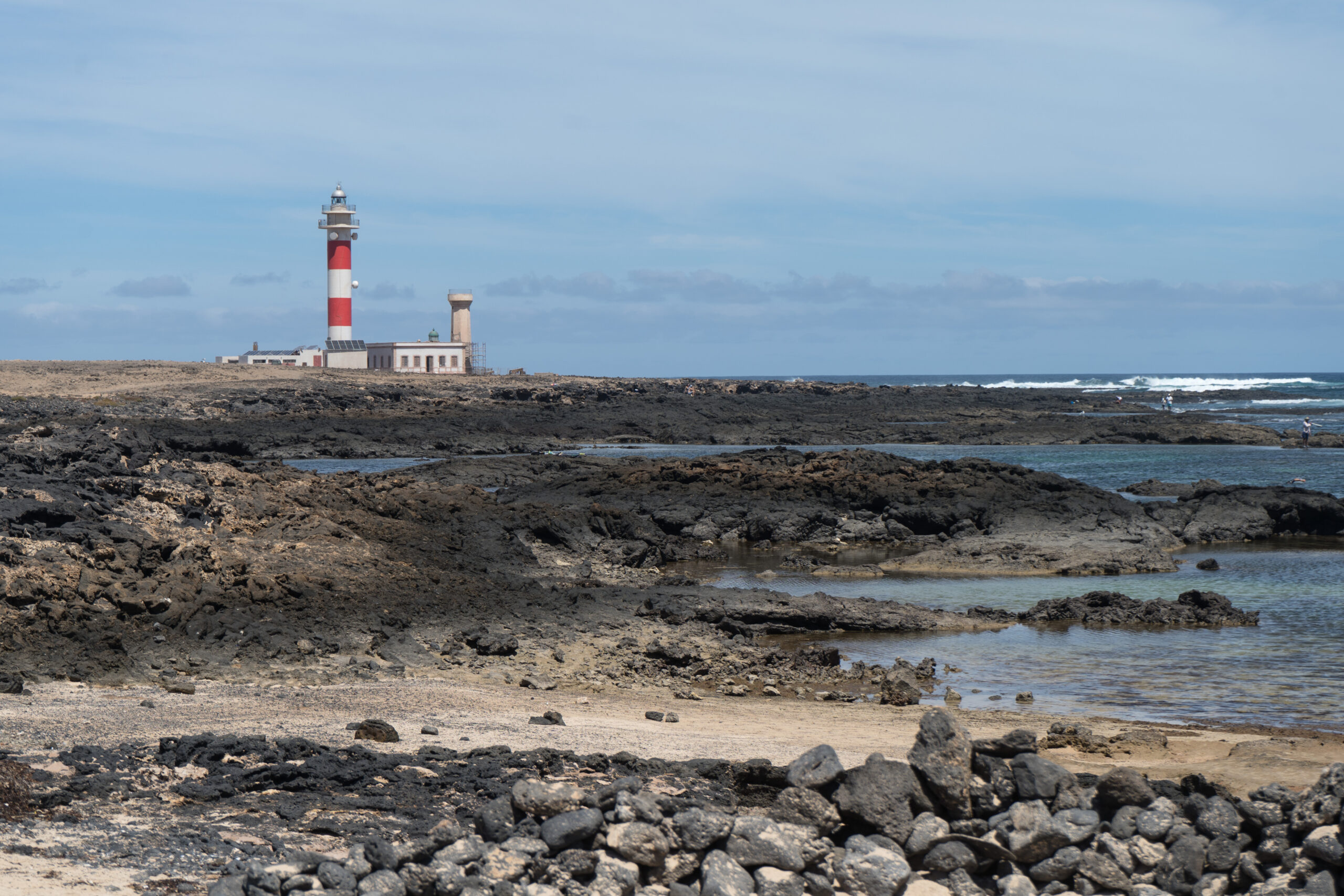 The Faro del Tostón lighthouse overlooks the rugged volcanic landscape of El Cotillo, with its distinctive red and white stripes visible across the rocky shoreline.