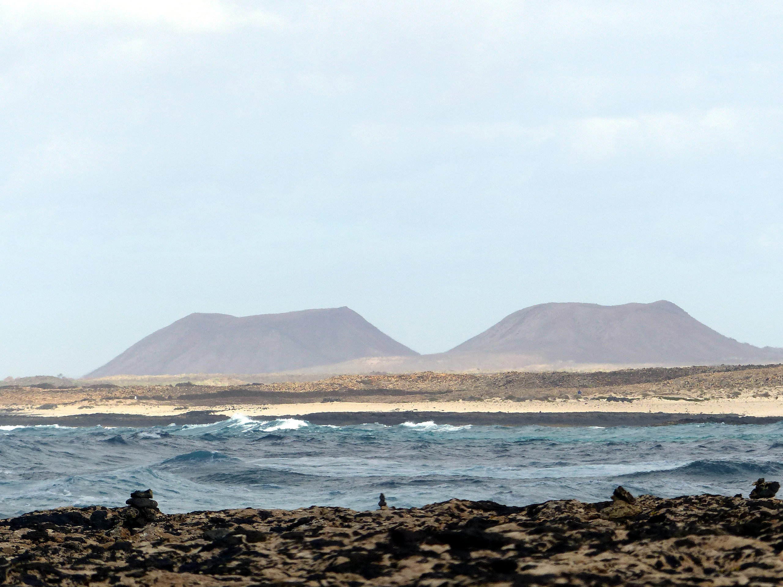 Volcanic peaks frame the coastal landscape near El Cotillo, with Atlantic waves rolling toward the dark volcanic rock shore.
