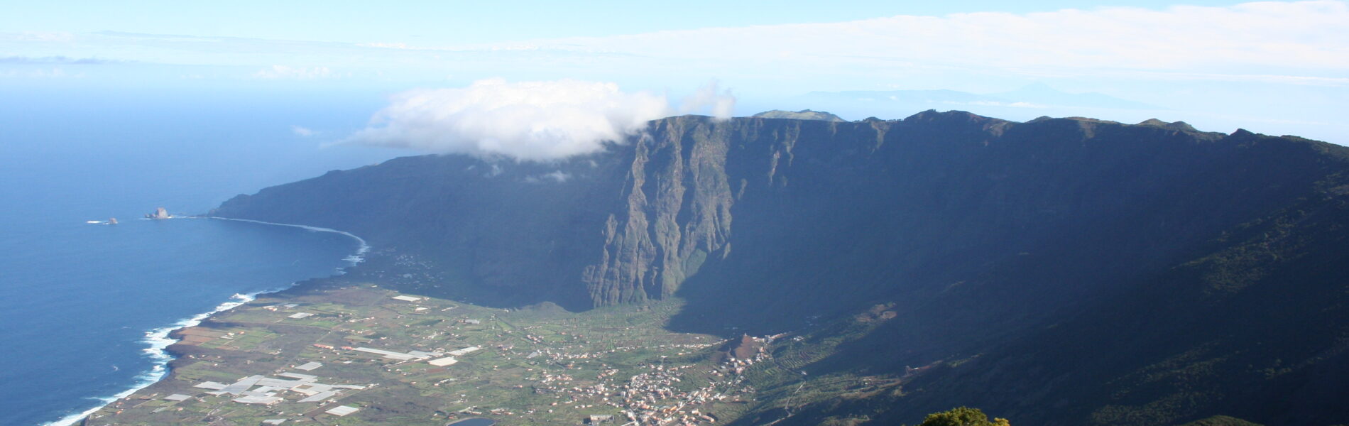 Aerial view of a steep-walled volcanic valley descending to a coastal plain with scattered buildings and the Atlantic Ocean beyond, under clear blue sky.