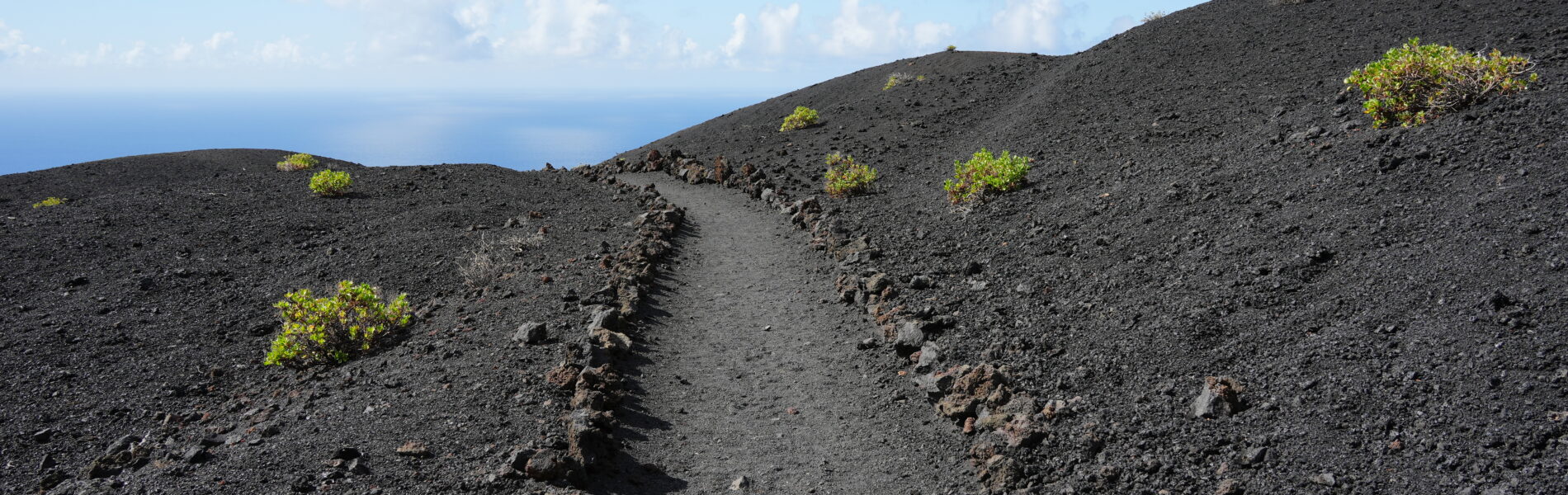 Hiking trail ascending through dark volcanic landscape with scattered green shrubs and blue ocean horizon