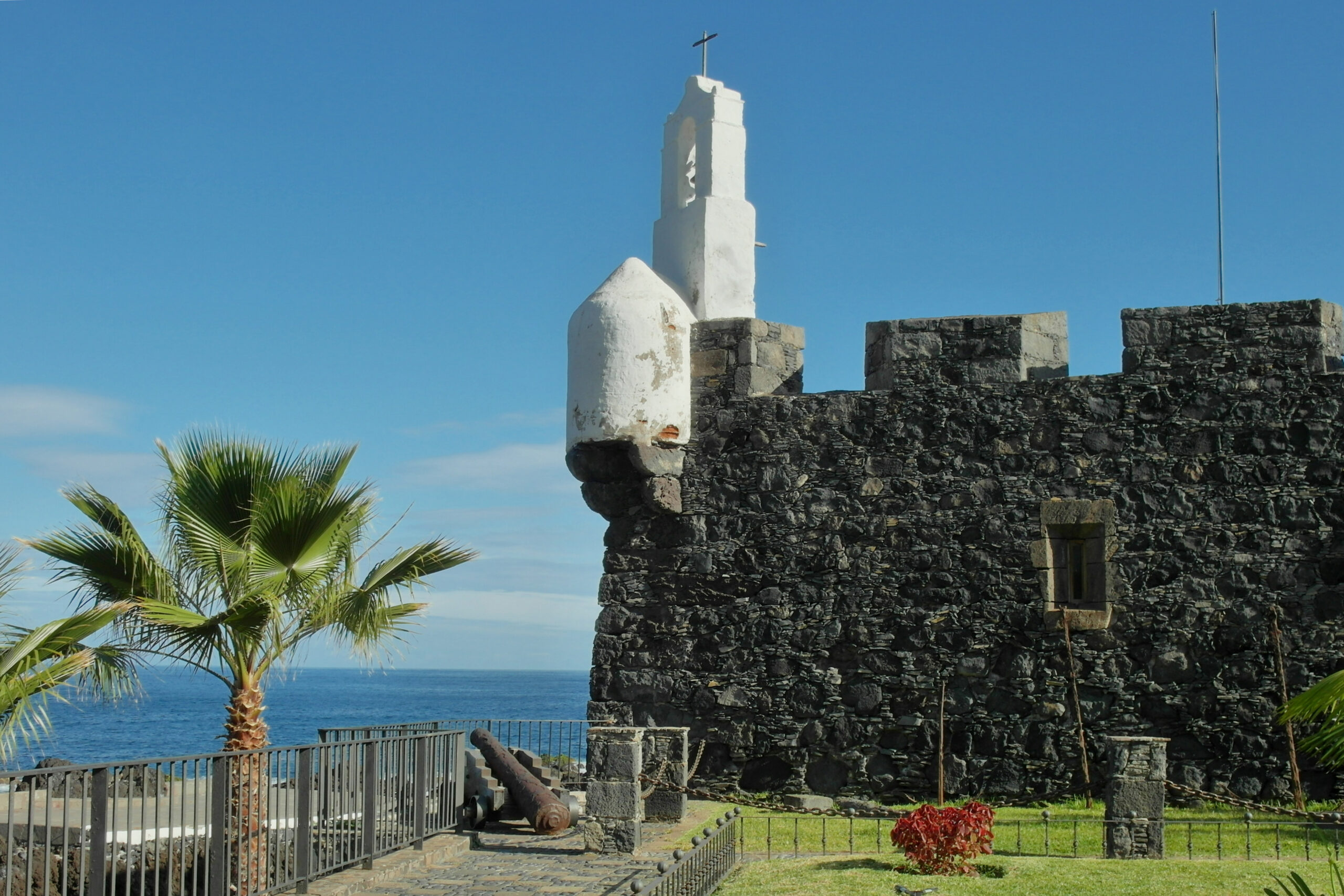Dark stone fortress with crenellated walls overlooks the sea, with a white chapel tower topped by a cross rising above the ramparts, palm trees and a cannon visible in the foreground.