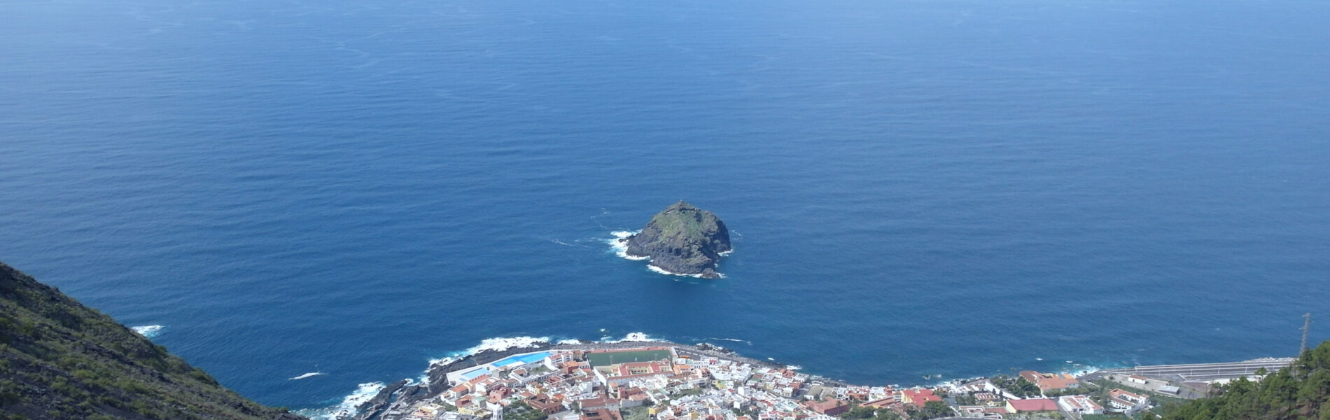 Aerial view of Garachico town with red-roofed buildings clustered on a peninsula, surrounded by deep blue ocean and a small rocky island offshore.