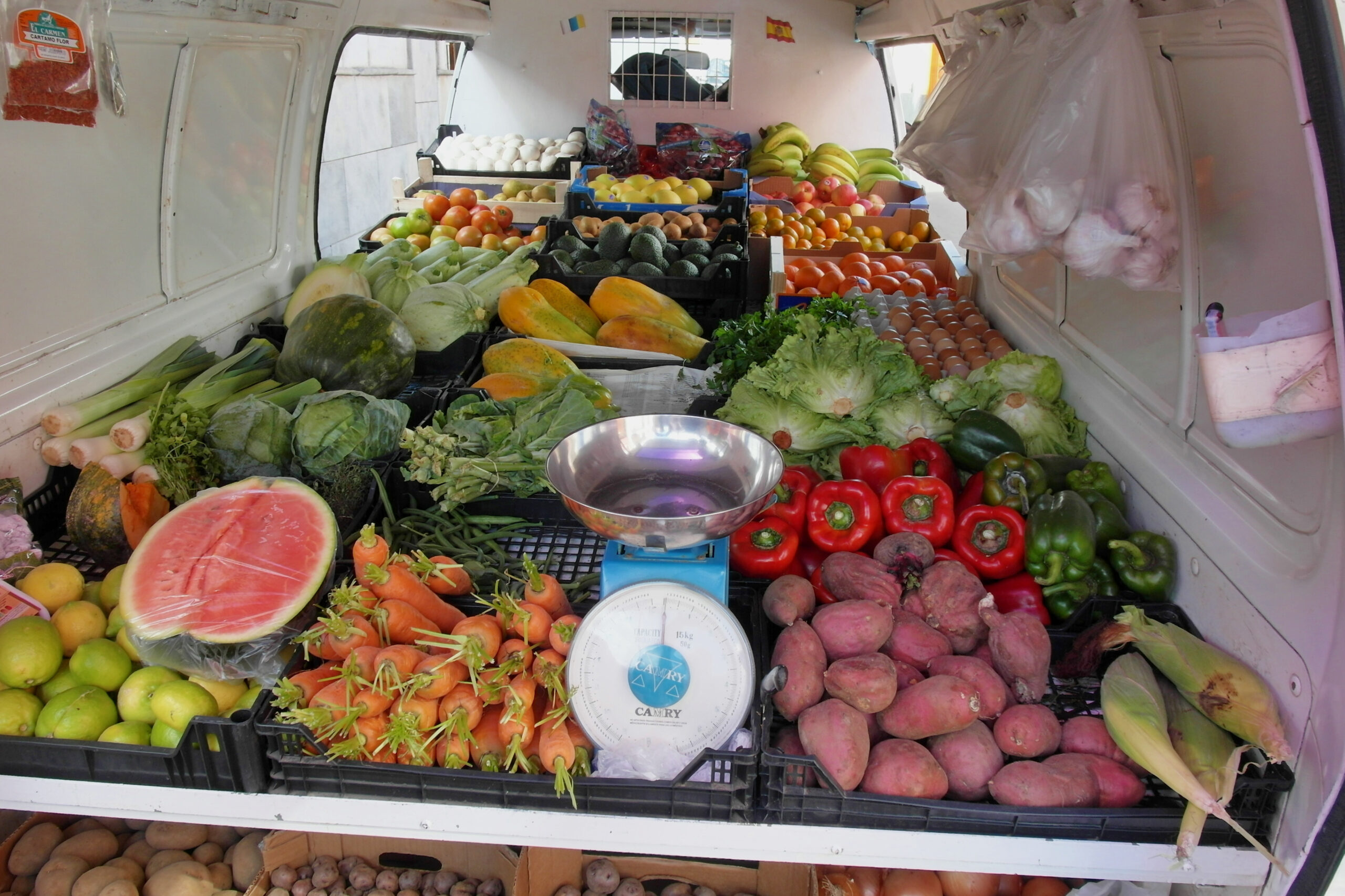 Interior of a white produce van loaded with fresh vegetables and fruits, including lettuce, peppers, tomatoes, onions, melons, and tropical fruit, with a metal scale in the center.