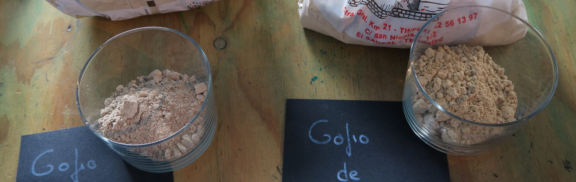 Two glass bowls of ground gofio flour on a wooden table with handwritten labels identifying them as red mullet and millet varieties, alongside packaged gofio products.