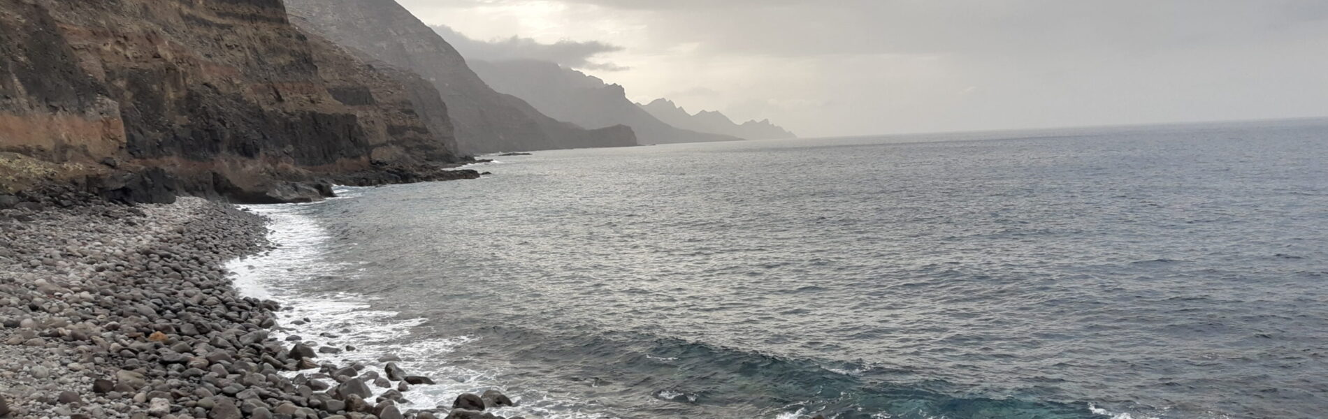 Rocky beach with pebbles and boulders meeting turquoise water, steep dark cliffs rising along the coastline under overcast sky