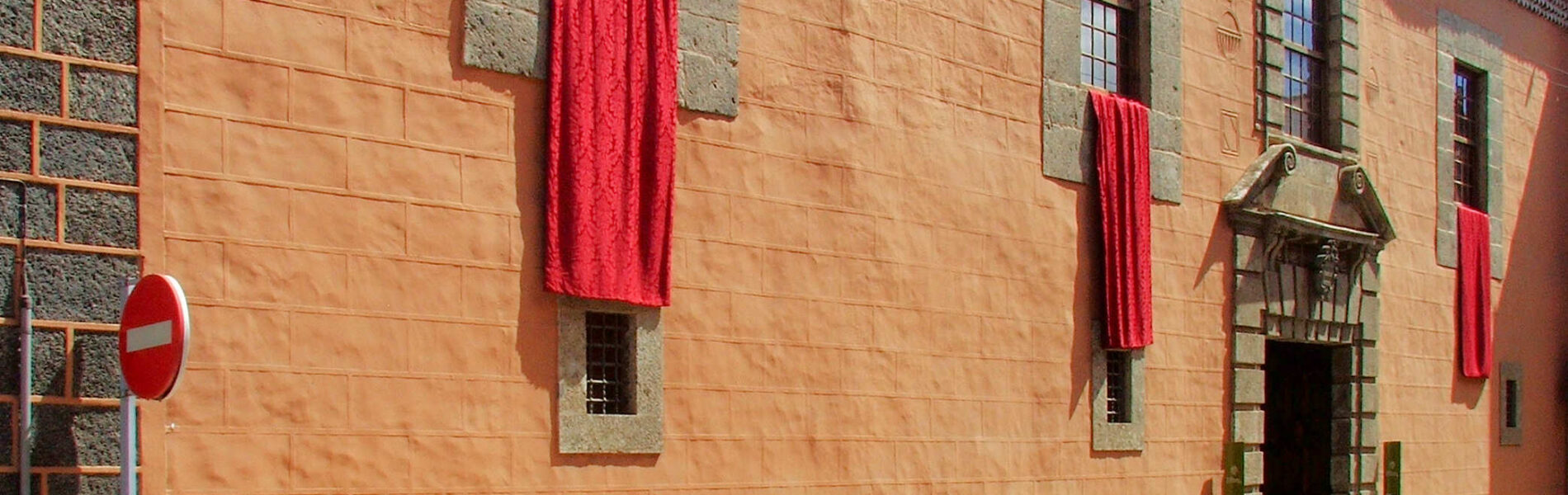 Terracotta-colored stone building with barred windows and decorative red fabric banners hanging from the facade, green-trimmed wooden doorway visible on right side.