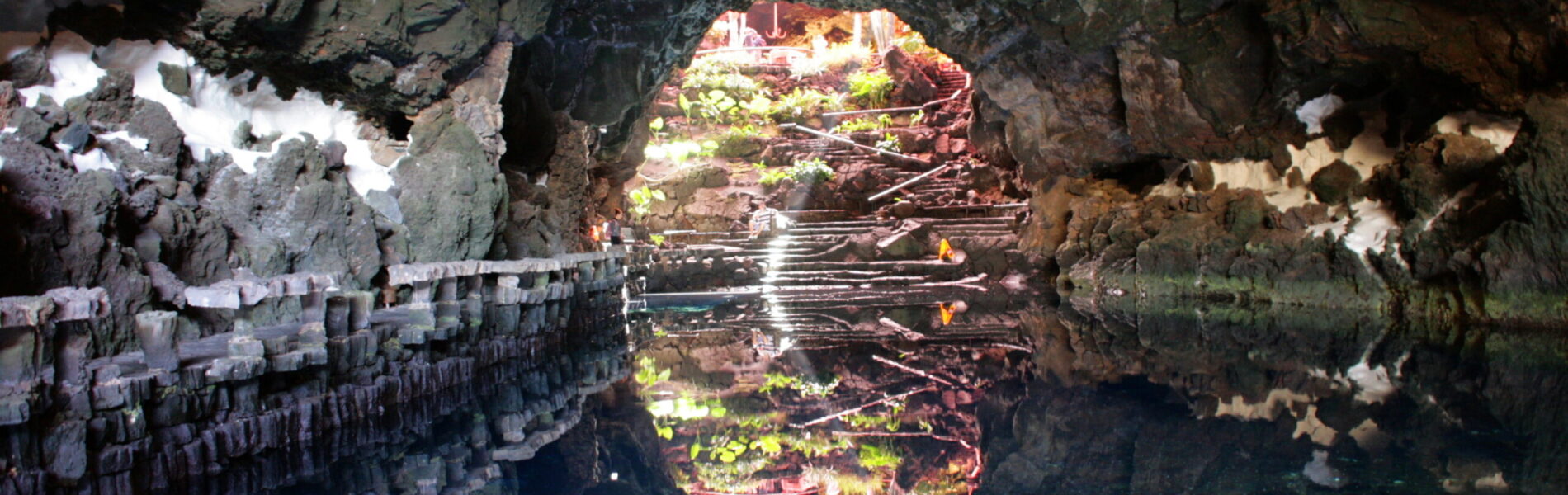 Underground cave chamber with still water reflecting volcanic rock walls and natural opening revealing lush vegetation outside