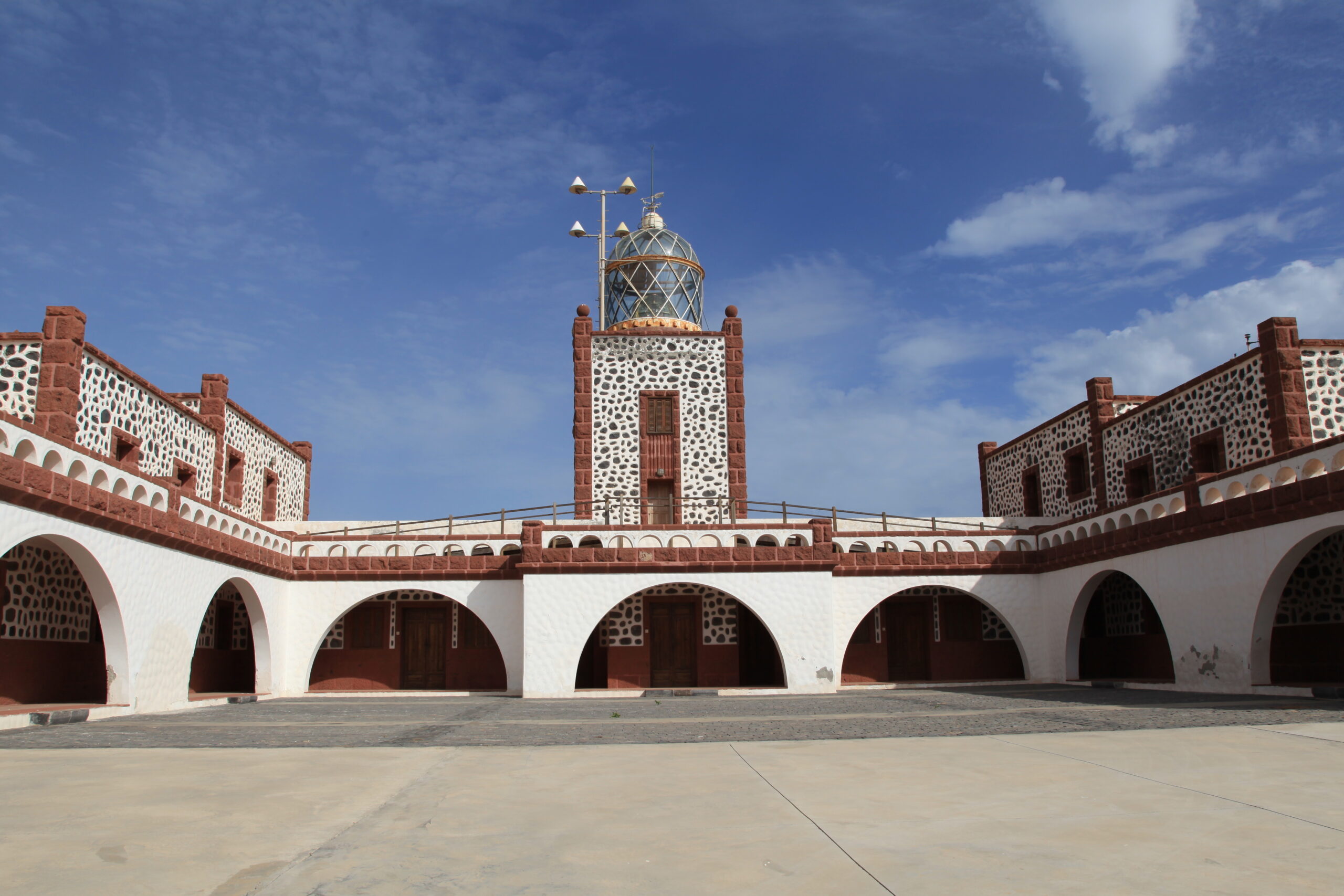 The striking red volcanic stone and white-painted arches of La Entallada Lighthouse frame a spacious courtyard, with the beacon tower rising prominently at its center.