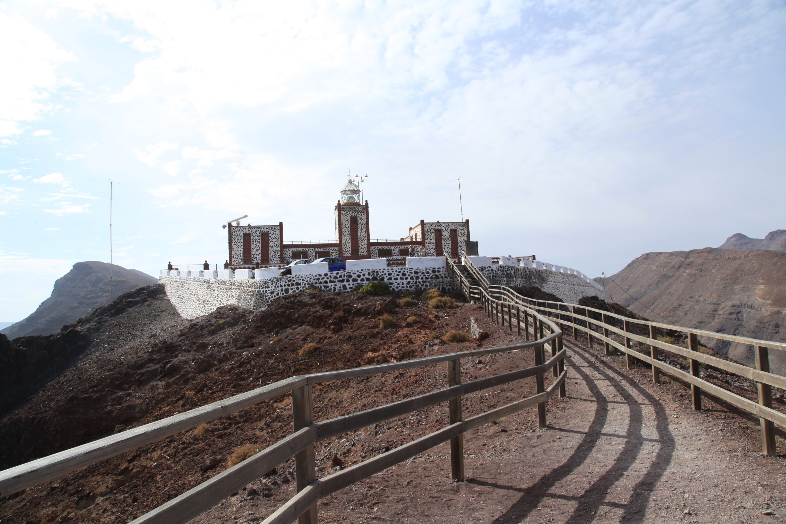 La Entallada Lighthouse stands on a dramatic volcanic ridge overlooking Las Playitas, accessible via a wooden walkway through the barren landscape.