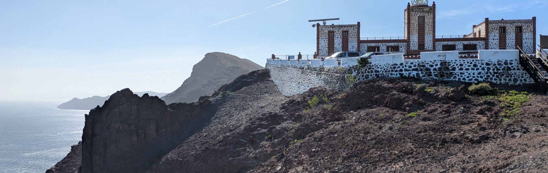 White lighthouse complex with towers on volcanic black rock hillside overlooking blue ocean and distant islands