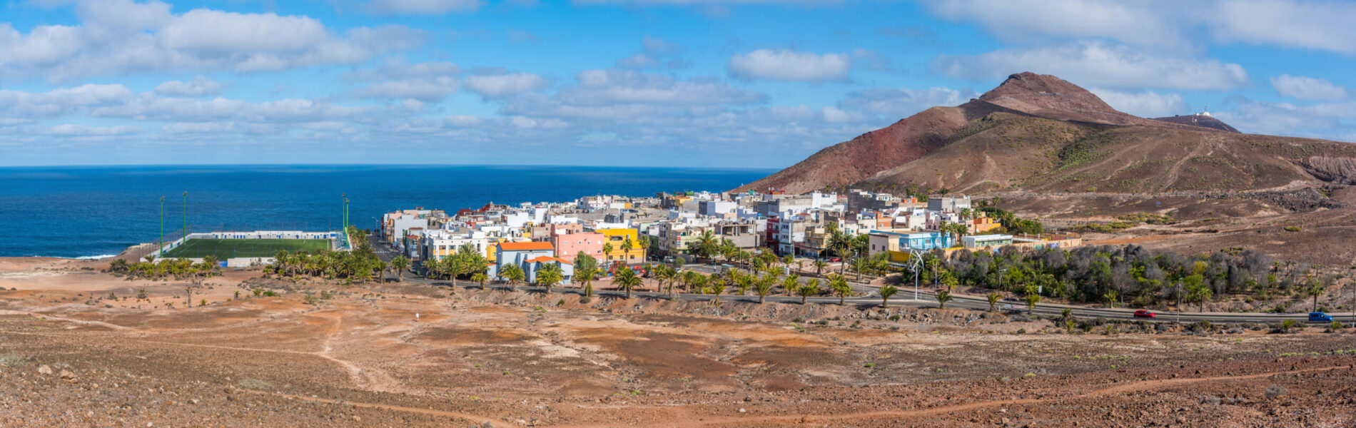 Colourful coastal neighbourhood set between volcanic slopes and the bright blue Atlantic under a partly cloudy sky.