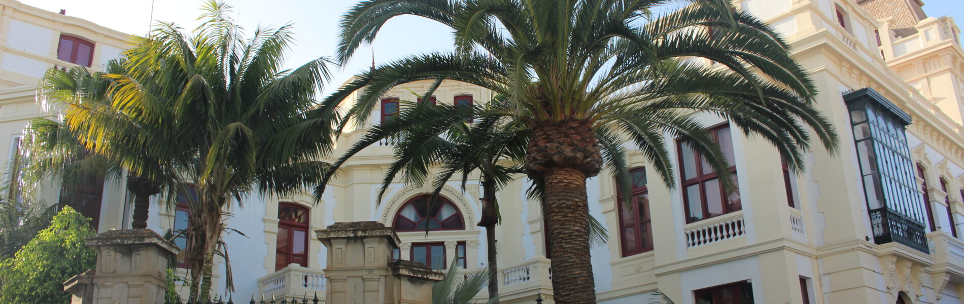 Cream-colored colonial building with red wooden doors and windows, fronted by tall palm trees and black iron gate, clear blue sky