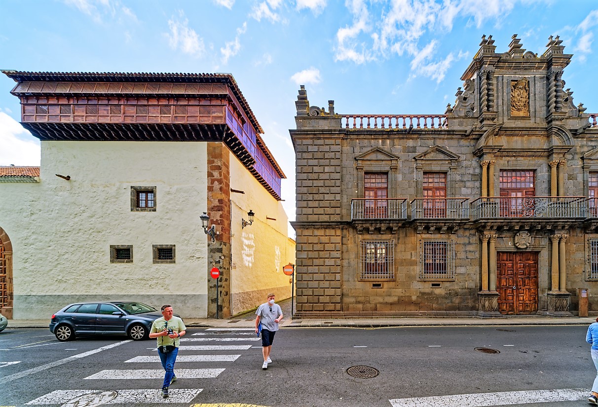 Colonial-era plaza with whitewashed and yellow buildings on the left, an ornate stone palace with red doors on the right, pedestrians crossing a zebra crossing, blue car parked nearby, clear sky above.
