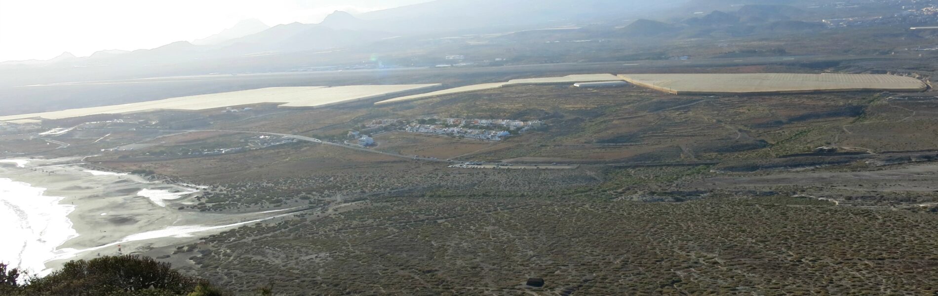Aerial view of a coastal landscape with salt marshes, dark volcanic terrain, a white sand beach, and mountains rising across the water under clear sky