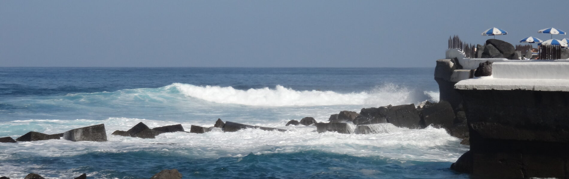 Rocky volcanic coastline with waves breaking against dark basalt rocks, white-domed structures visible on a platform at right, clear blue sky and ocean horizon.
