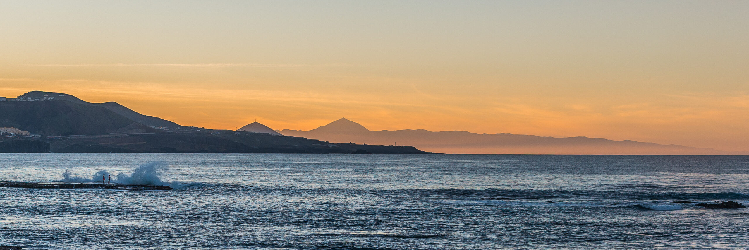 Las Canteras Beach glows in warm evening light, with volcanic peaks visible across the water at sunset.