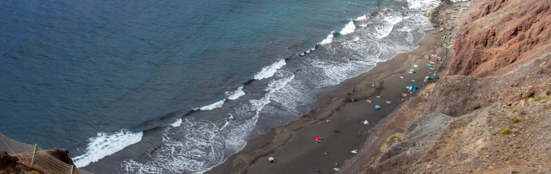 Aerial view of a black sand beach with white-capped waves, surrounded by steep reddish-brown cliffs and sparse vegetation, with colorful beach umbrellas dotting the sand.