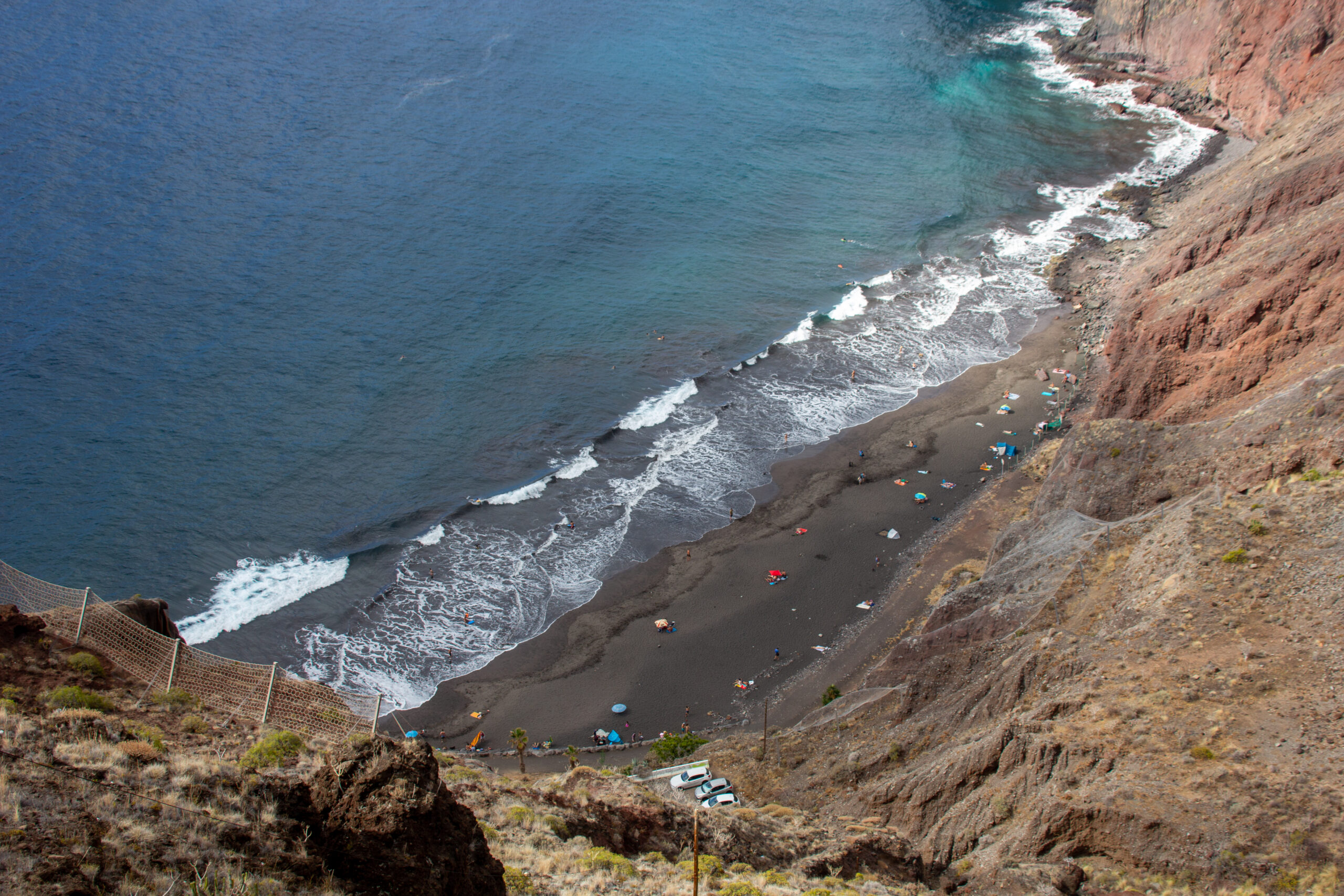 Aerial view of a black sand beach with white-capped waves, surrounded by steep reddish-brown cliffs and sparse vegetation, with colorful beach umbrellas dotting the sand.