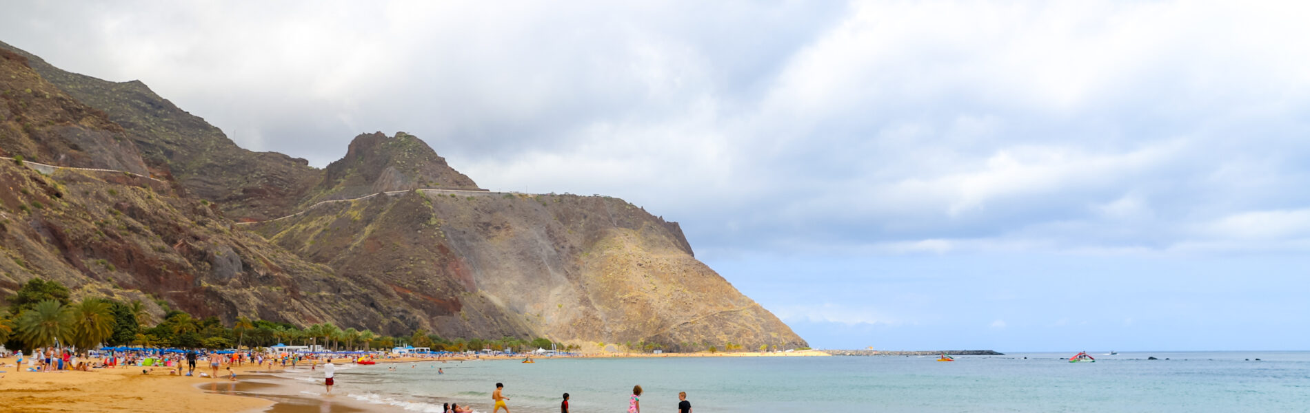 Golden sandy beach with swimmers in clear turquoise water, backed by a dramatic volcanic cliff with striations of red and yellow rock under partly cloudy sky.