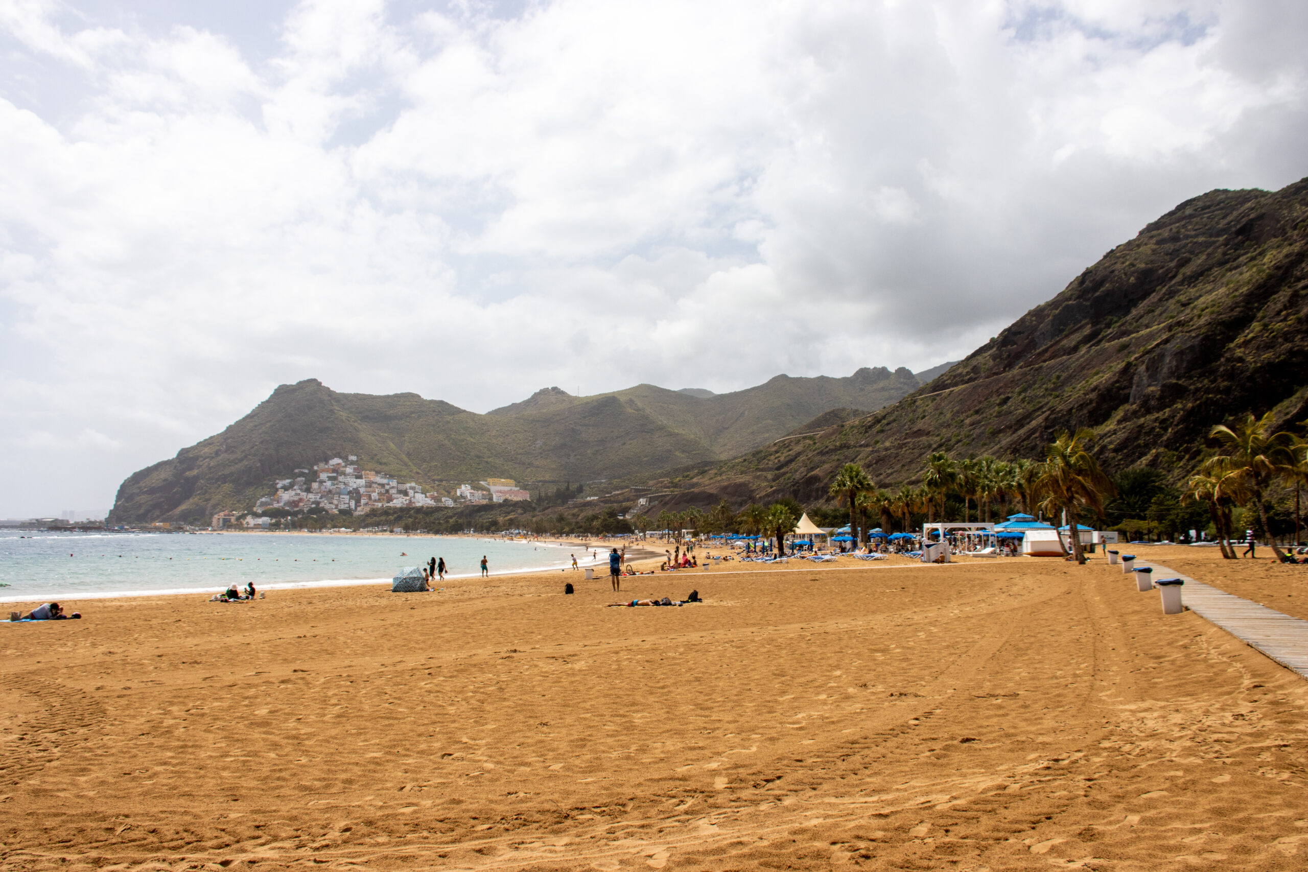 Golden sandy beach with swimmers and sunbathers, backed by palm trees and dramatic green volcanic mountains under partly cloudy sky