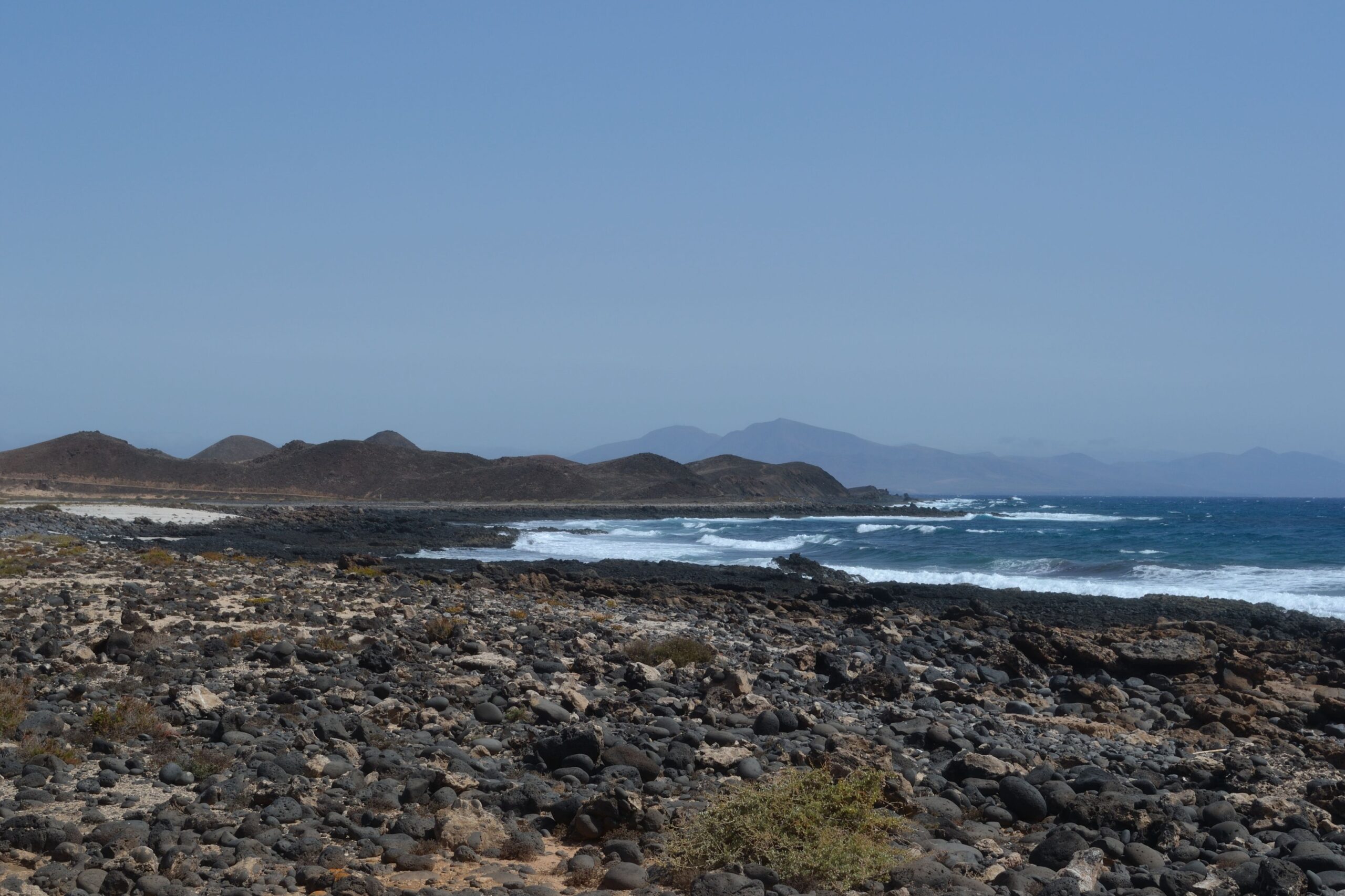 The rugged volcanic landscape of Lobos Island features dramatic dark lava rock formations along its coastline, with views across the strait toward Fuerteventura's distant peaks.