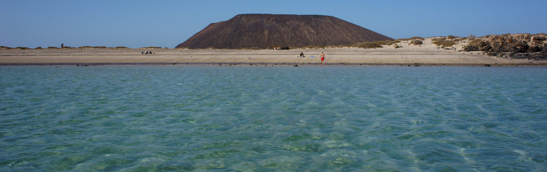 Clear turquoise water with sandy beach and a distinctive dark volcanic cone rising behind it under blue sky