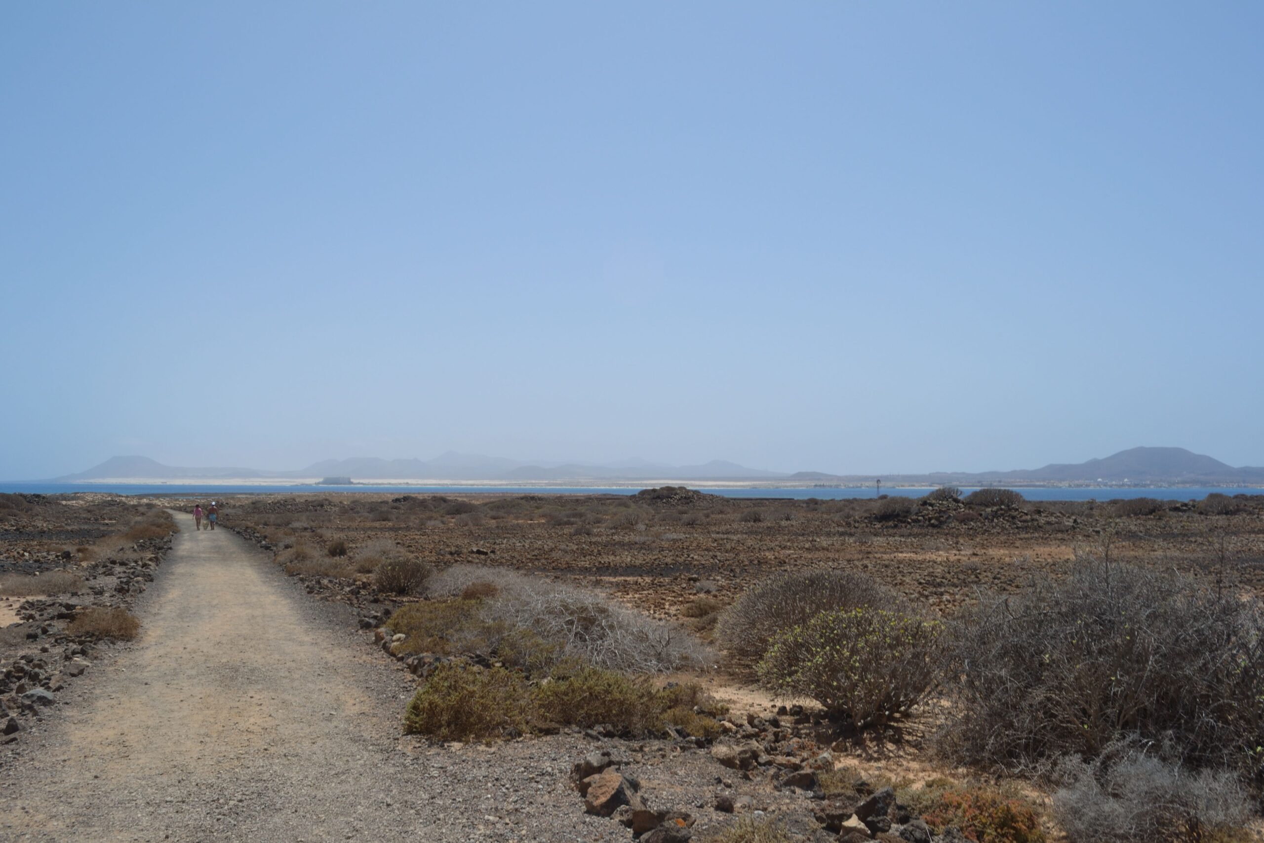 A walking trail traverses the rocky, sparsely vegetated landscape of Lobos Island, with views across to the surrounding waters and distant peaks.