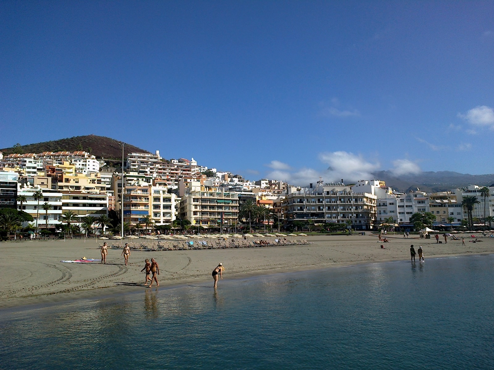 Beachgoers swim and sunbathe on sandy beach with colorful apartment buildings and hotels rising on hillside under clear blue sky