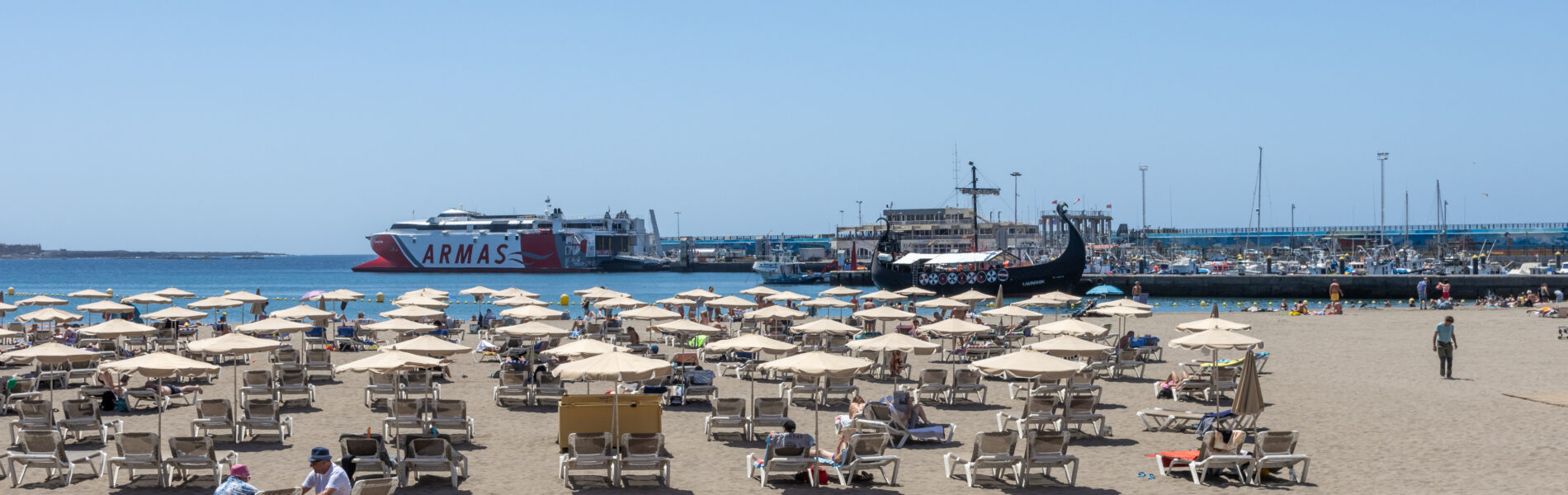 Sandy beach with dozens of cream-colored umbrellas and loungers, ferries and sailboats in blue harbor water beyond, clear sky