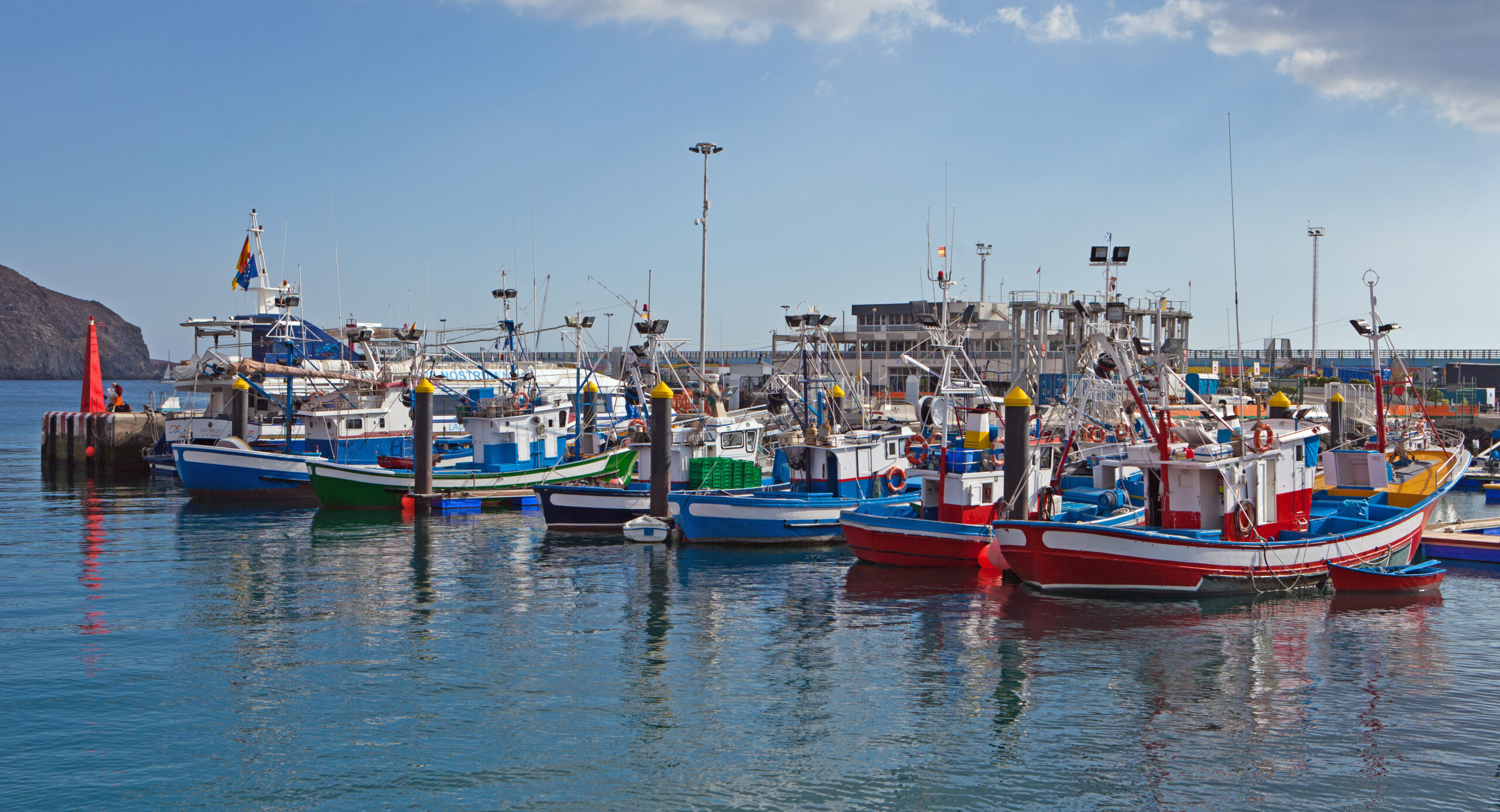 Colorful fishing boats moored in a calm harbor with blue and red hulls, masts and rigging visible against clear sky and rocky coastline.