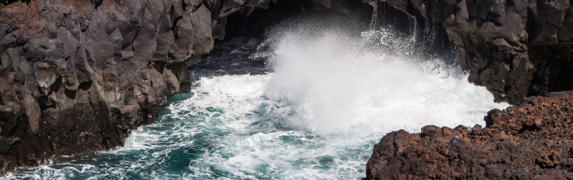Turquoise waves crash against dark volcanic rock formations in a cove, sending white spray high into the air.