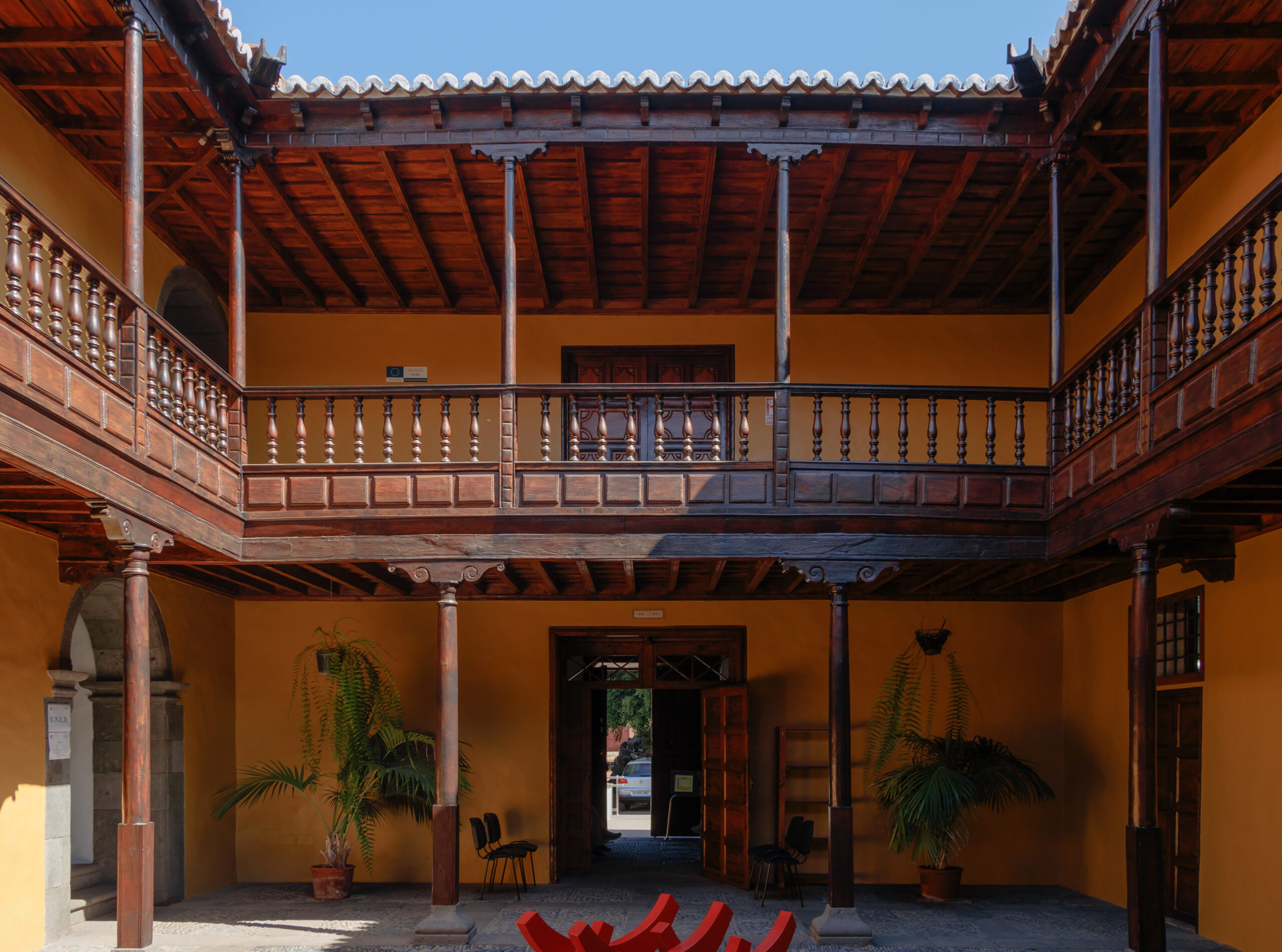 A traditional colonial-style courtyard features vibrant ochre walls, intricately carved wooden galleries, and period architectural details characteristic of historic buildings in Los Llanos.