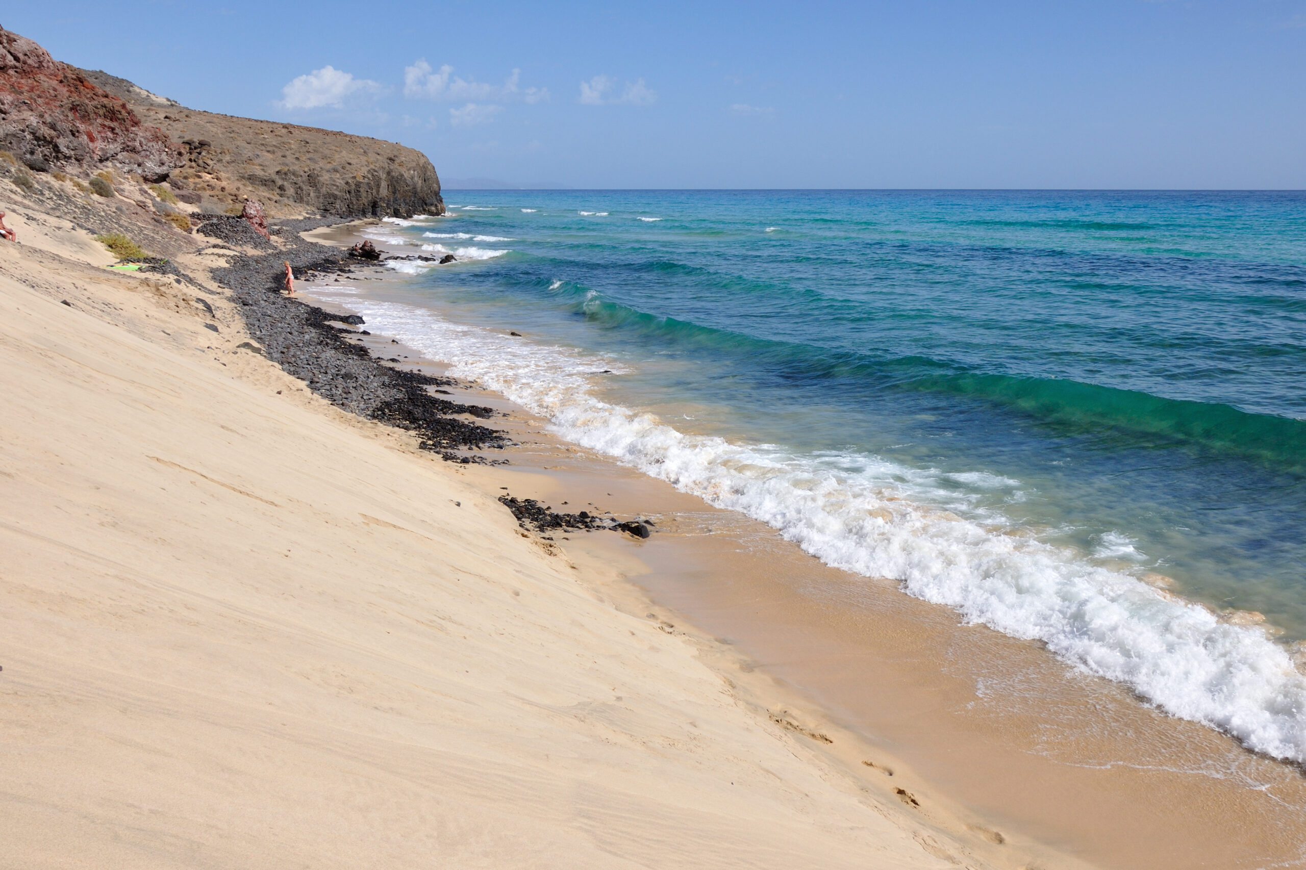 Mal Nombre Beach stretches along the Jandía peninsula with golden sand, rolling surf, and dramatic coastal cliffs rising above the shoreline.