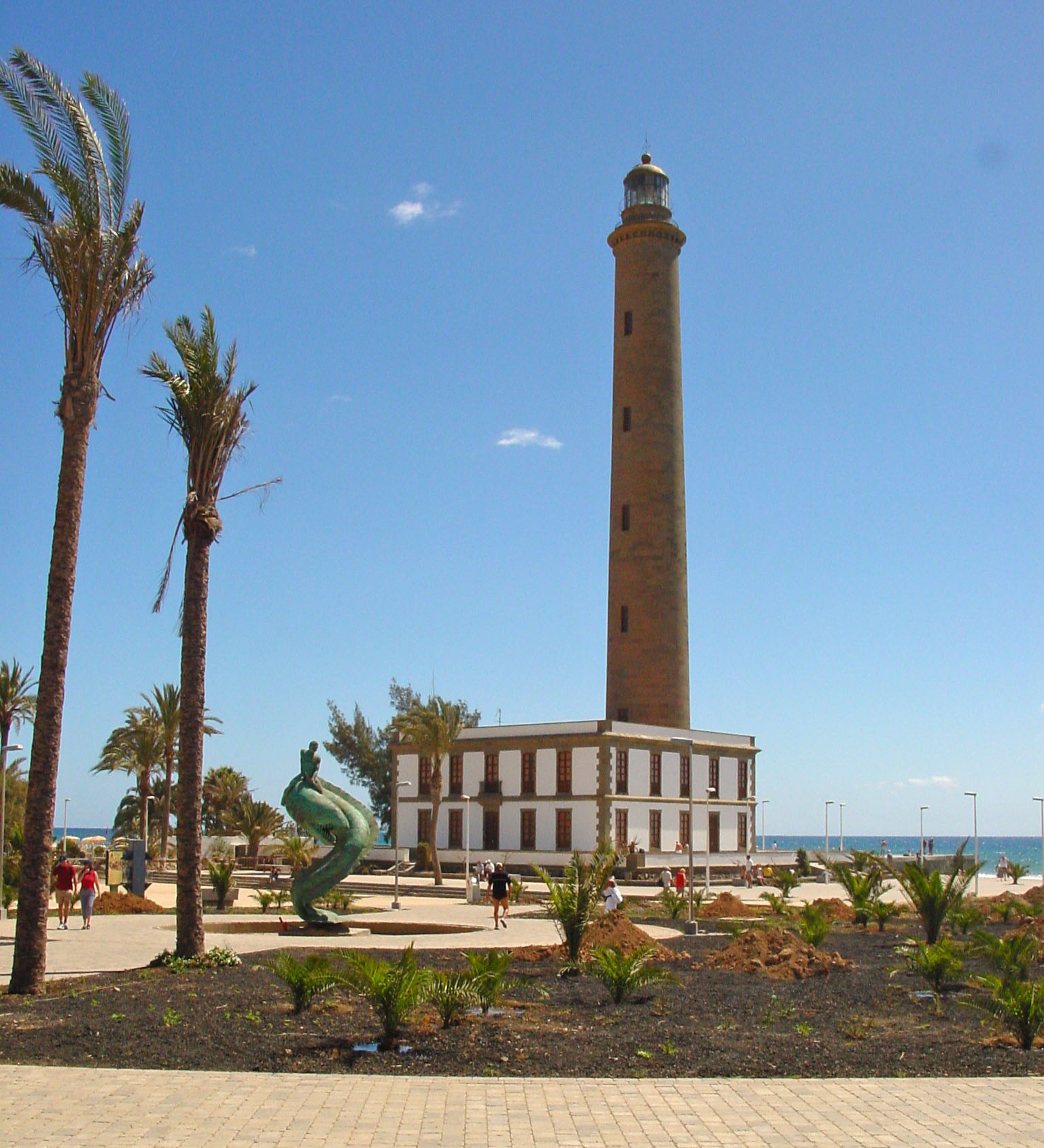 The historic Maspalomas Lighthouse stands prominently above the promenade, framed by palms and the Atlantic beyond.