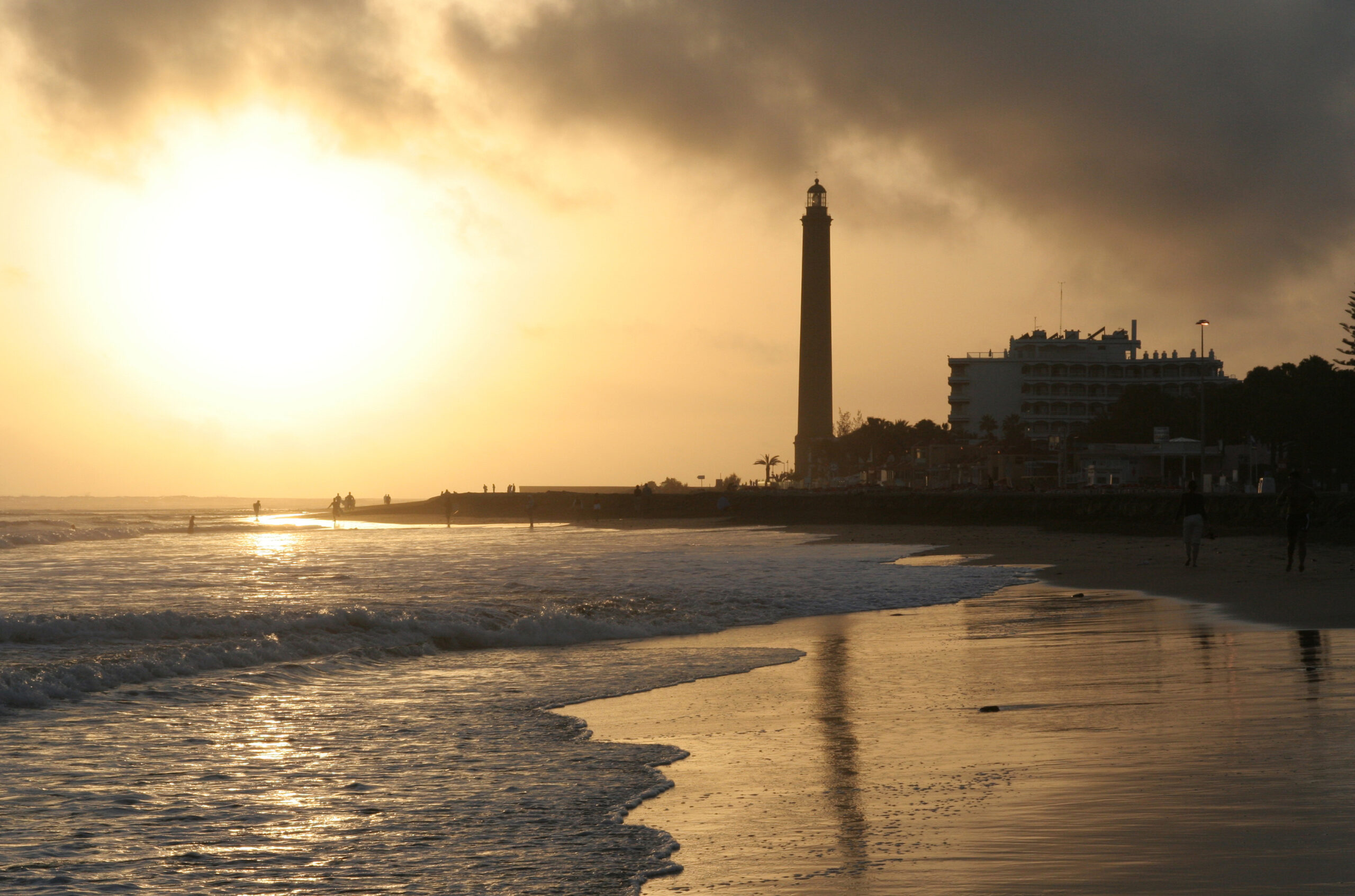 The Maspalomas Lighthouse stands sentinel at sunset, with gentle waves lapping the sandy shore below.