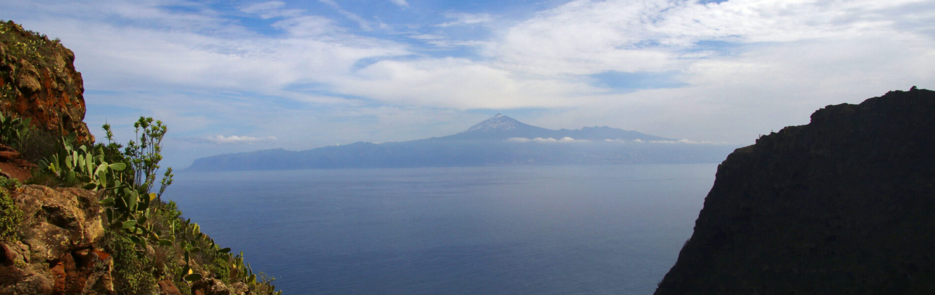 Rocky cliffsides with succulent vegetation frame a distant volcanic peak across a deep blue ocean under partly cloudy sky