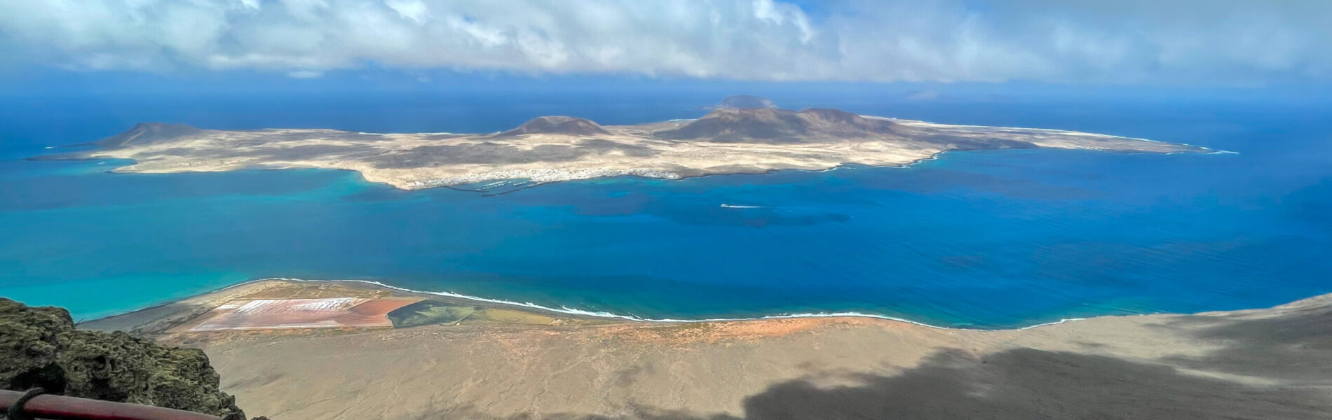 Red metal railing on dark volcanic rock overlooking turquoise ocean, golden islands, and blue sky with white clouds